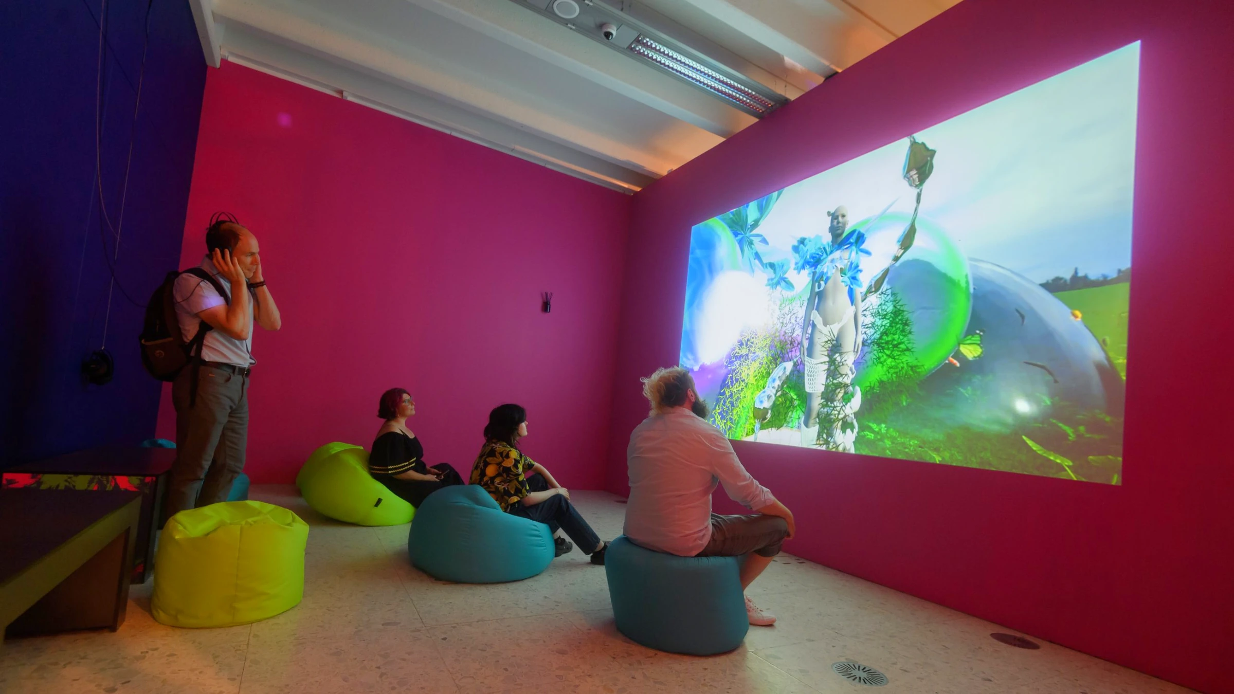 Visitors seated on bean bags in front of the projected Wing Possibility film in a magenta gallery room, one visitor listening on headphones.